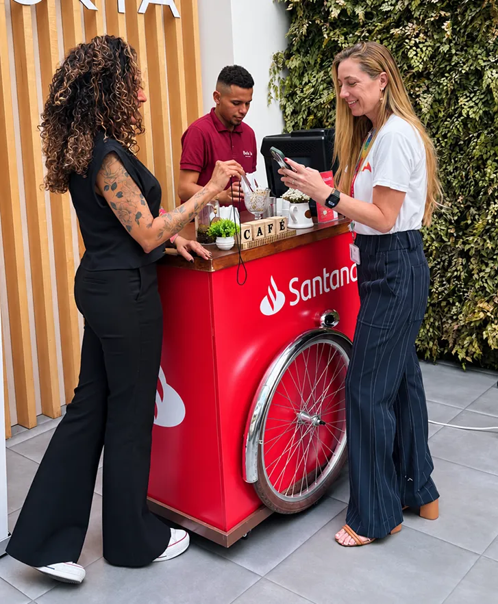 Convidadas conversando no carrinho de café com a identidade visual do Santander enquanto o barista prepara o café